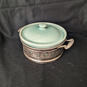 May include: A light blue ceramic casserole dish with a lid and a silver metal stand with decorative cutouts.