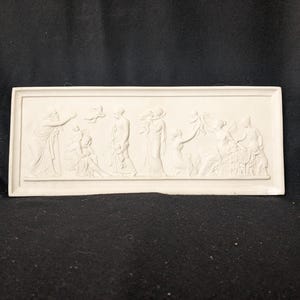 Puede incluir: Una escultura rectangular en relieve de color blanquecino que representa figuras clásicas con túnicas fluidas. El bajorrelieve detallado muestra múltiples figuras en varias poses, algunas sosteniendo objetos. El tema general sugiere una escena histórica o mitológica.
