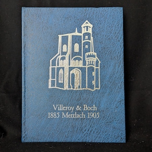 May include: A blue book with a silver embossed illustration of a building. The text "Villeroy & Boch 1885 Mettlach 1905" is printed below the illustration. The book has a textured cover.