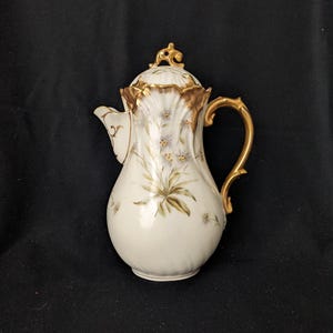 Peut inclure: Cafetière en porcelaine blanche avec une anse, un bec et un couvercle dorés. Le pot est décoré de motifs floraux dans des tons de vert et de violet. Le couvercle est surmonté d'un élément décoratif doré. Le pot est posé sur un fond noir.