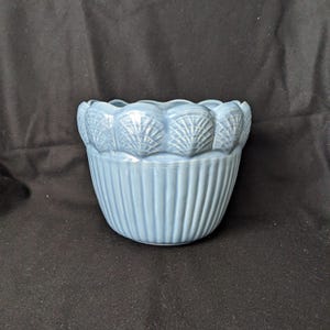 以下が含まれることがあります： 縁に貝殻のデザインが施された、水色のセラミックボウル。ボウルの側面には垂直方向のリブのディテールがあります。ボウルは水色で、装飾目的で使用される可能性があります。