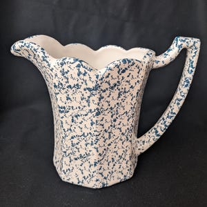 Puede incluir: Una jarra de cerámica blanca con un esmalte moteado azul. La jarra tiene un borde festoneado y un asa.