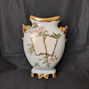 Puede incluir: Un jarrón de cerámica azul claro con detalles dorados, adornado con diseños florales rosas y dos marcos rectangulares en blanco. El jarrón tiene asas doradas decorativas y una base, perfecto para la decoración del hogar.