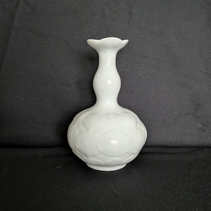 Puede incluir: Un jarrón de cerámica blanca con una base bulbosa y un cuello delgado. La base presenta un patrón floral en relieve repetido. La parte superior del jarrón tiene una abertura acampanada en forma de pétalo. El jarrón mide unos 18 cm de alto.