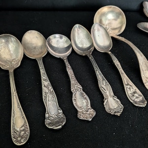 Puede incluir: Una colección de seis cucharas de metal plateado con patrones florales y de hojas en relieve en los mangos. El conjunto incluye cinco cucharas con cuencos ovalados y un cucharón más grande con cuenco redondo, todos con detalles decorativos.
