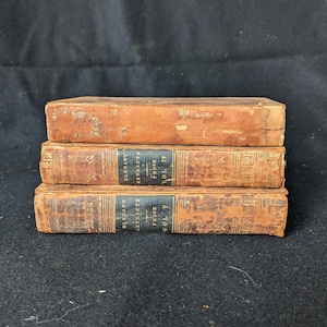 May include: Three antique books stacked on top of each other. The books have worn, brown leather covers. The spines of the bottom two books have black labels with gold lettering that reads "ELEGANT EXTRACTS PROSE VOL IV" and "ELEGANT EXTRACTS PROSE VOL V".