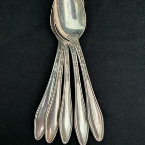May include: A set of four silver-toned metal spoons arranged in a fan pattern. Each spoon features a polished oval bowl and a slender handle decorated with an embossed diamond and leaf pattern that extends from the neck down the center of the stem.