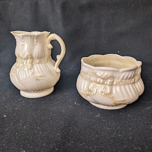 Puede incluir: Un juego de jarra de crema y azucarero de porcelana de color crema con un delicado diseño de lazo en relieve. El juego presenta un borde festoneado y un glaseado amarillo sutil.