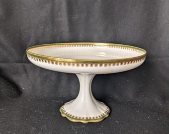 Puede incluir: Soporte para pasteles de porcelana blanca con ribete dorado y verde. El soporte tiene un plato ancho y poco profundo y una base acanalada. El borde del plato y la base están decorados con un patrón dorado y verde.