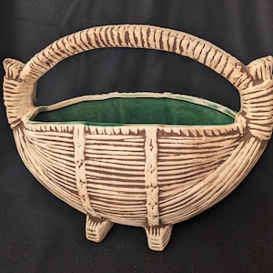 Puede incluir: Un macetero de cerámica con forma de cesta y asa. El macetero es blanco con detalles marrones y tiene un interior verde esmaltado.