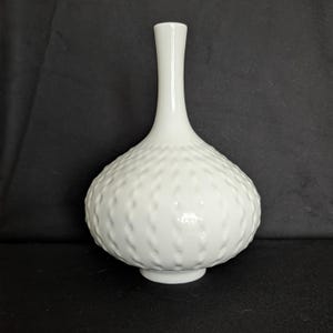 Puede incluir: Un jarrón de cerámica blanca con un cuerpo bulboso y un cuello largo y delgado. El cuerpo presenta un patrón texturizado y punteado. El jarrón mide aproximadamente 25 cm de alto y tiene un acabado brillante. Es un artículo decorativo.