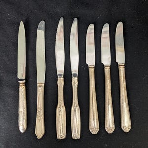 May include: A collection of eight silver knives with varying blade shapes and handle designs. Some knives have serrated edges, while others have smooth blades. The handles feature ornate patterns, including floral and geometric motifs. The knives are arranged on a dark surface.