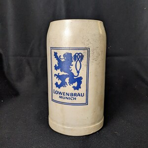 May include: A vintage, off-white ceramic beer stein with a blue logo featuring a lion and the text "Löwenbräu Munich." The stein has a cylindrical shape and a slightly textured surface. The logo is in a rectangular frame.