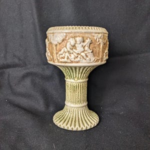 Puede incluir: Una jardinera de pedestal de cerámica con un diseño en relieve detallado. La jardinera presenta una base verde claro y una sección superior beige con una escena clásica de figuras. El diseño es una pieza decorativa y detallada.