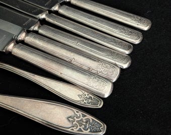 May include: A collection of silver-toned metal knives and spoons featuring ornate, embossed floral and urn-shaped detailing on the handles. The cutlery set displays a polished metallic finish with intricate decorative patterns at the base.