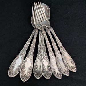 May include: A set of silver-toned metal forks and spoons featuring an embossed grape and vine pattern on the handles. The utensils are arranged in a fan-like cluster, showcasing the detailed metallic texture and the ornate floral motif.