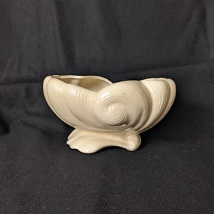 May include: Cream-coloured ceramic seashell-shaped planter. The planter has a wide opening and a textured, swirling design on the sides, with a base that resembles a shell. Suitable for small plants or decorative items.