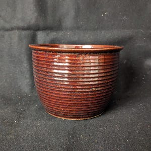 May include: A small, round, ceramic pot with a dark reddish-brown glaze. The pot has horizontal, ridged detailing around the exterior and a smooth rim. The pot is likely used for planting small plants or as a decorative item.