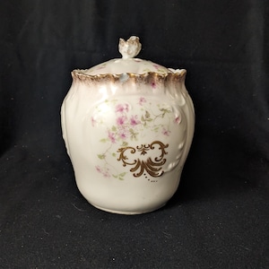 May include: A white porcelain jar with a lid, featuring pink floral patterns and gold accents. The jar has a rounded shape with a decorative gold trim around the rim and a small floral knob on the lid. The design includes gold filigree details.