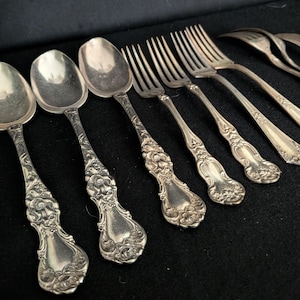 May include: A collection of silver-toned metal flatware including two spoons and three forks arranged in a row. Each piece features an ornate, embossed floral pattern on the handle, with a polished finish and detailed scrollwork at the base.