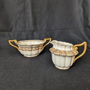 May include: A white porcelain sugar bowl and creamer set. The sugar bowl has two gold handles and a decorative band with pink, gold, and black accents. The creamer has a spout and a gold handle, with a similar decorative band.
