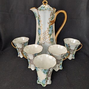 May include: A vintage porcelain coffee set comprising a tall, ornate coffee pot and five matching cups. The set is white with gold accents and delicate blue floral patterns. The cups have gold handles and a lattice design.