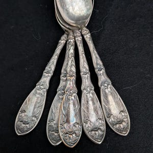 May include: A set of four silver-toned metal spoons featuring intricate embossed floral patterns along the handles. The spoons have a tarnished finish with detailed engravings of flowers and swirling vines extending from the base to the neck.