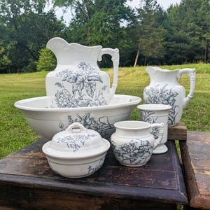 Op de afbeelding: Een verzameling wit keramisch servies met een blauw bloemmotief. De set bevat een grote kan, een waskom, een kleine kan, een afgedekte kom en een kleine beker. De stukken zijn op een houten oppervlak geplaatst.