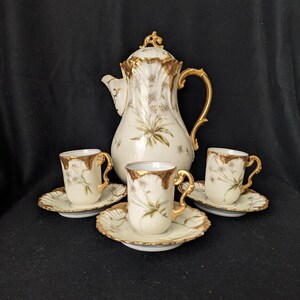 Könnte beinhalten: Ein Vintage-Porzellan-Kaffeeservice mit einer cremefarbenen Kaffeekanne und drei passenden Tassen und Untertassen. Das Set ist mit goldenen Verzierungen und zarten Blumenmustern verziert. Die Kaffeekanne hat einen dekorativen Deckel und Griff.