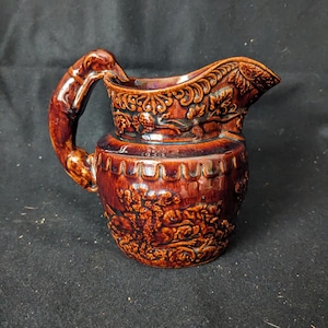 May include: A brown ceramic pitcher with a detailed relief design. The pitcher features a curved handle and a spout. The relief depicts floral and landscape scenes. The pitcher is a decorative item.