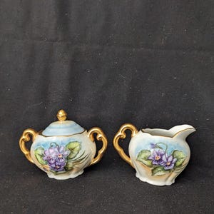May include: A porcelain sugar bowl with a lid and a matching creamer pitcher. Both pieces feature hand-painted purple violet flowers with green leaves against a light blue background, accented by ornate gold handles and gold trim details.