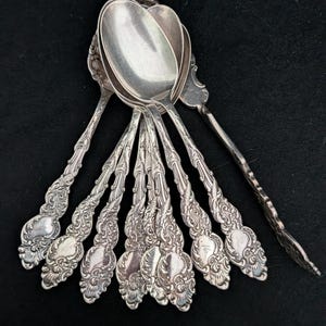 May include: A collection of seven silver-toned metal spoons arranged in a fan shape. Each spoon features an ornate, embossed handle with intricate scrollwork and floral-inspired detailing, leading to a smooth, polished, oval-shaped bowl.