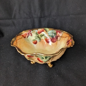 Könnte beinhalten: Eine kleine, antike Schale mit Füßen, einem gewellten Rand und goldenem Besatz. Die Schale zeigt ein handgemaltes Design von Erdbeeren und Blättern in Rot-, Grün- und Brauntönen auf cremefarbenem Hintergrund.