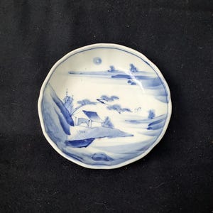 May include: A small, white ceramic dish with a blue landscape design. The scene depicts a house, trees, and mountains. The dish has a wavy edge and is likely used for serving small portions of food or as a decorative item.