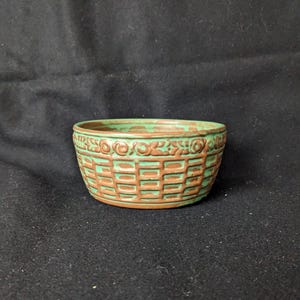 Puede incluir: Un pequeño cuenco redondo de cerámica con un diseño de tejido de cesta. El cuenco está pintado en tonos verdes y marrones. El borde del cuenco tiene un patrón decorativo. El cuenco probablemente se utiliza para servir o exhibir.