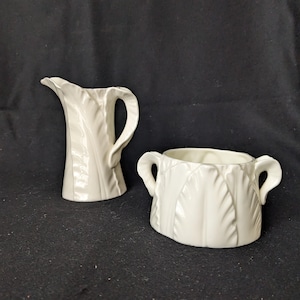 May include: A white ceramic creamer and sugar bowl set. The creamer has a curved handle and a spout. The sugar bowl has two handles. Both pieces have a textured leaf design. The set is on a dark background.