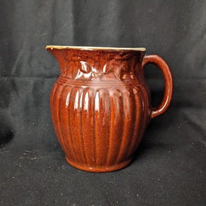 Può includere: Una brocca in ceramica marrone con un design a coste e un manico. La brocca ha un bordo leggermente svasato con un bordo color crema. Accenti decorativi a forma di foglia sono vicino alla parte superiore. La brocca viene probabilmente utilizzata per servire bevande.