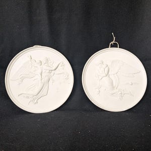 Peut inclure: Deux plaques murales rondes blanches avec des motifs d'anges en relief. L'une représente un ange avec un enfant, l'autre un ange agenouillé. Elles ont une boucle pour être accrochées.