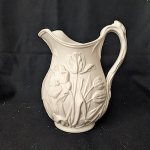 May include: A white ceramic pitcher with a floral relief design. The pitcher features a curved handle and a spout. The embossed flowers include tulips and other blossoms. The pitcher is likely used for serving beverages or as a decorative item.