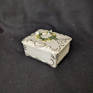 May include: A small, white porcelain trinket box with a square shape. The lid is decorated with a cluster of white and yellow roses. Gold trim accents the edges and decorative patterns. A charming decorative piece.