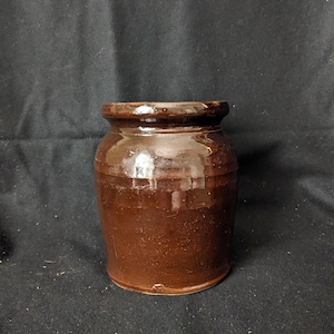 Puede incluir: Un tarro de cerámica marrón vidriado, con un cuerpo redondeado y un borde ligeramente ensanchado. El tarro tiene un acabado brillante y parece ser vintage. El color marrón oscuro y el diseño simple sugieren que podría usarse para almacenamiento o decoración.