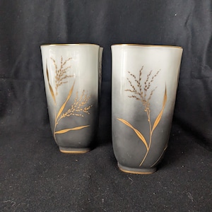 May include: Two tall, elegant glasses with a gradient design, transitioning from white to dark gray. Gold-colored botanical illustrations adorn the front. The rims and bases are trimmed with gold. These glasses are ideal for serving beverages.