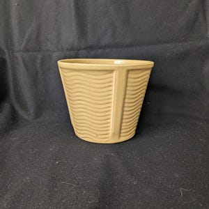Puede incluir: Una maceta de cerámica beige con un diseño ondulado y texturado. La maceta tiene una abertura redonda ancha y una base ligeramente cónica.
