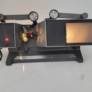 Puede incluir: Un editor de películas negro y plateado con una pantalla incorporada. La pantalla está iluminada y el editor tiene un panel de control con el texto "8 mm FILM EDITOR" en él.