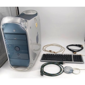 Op de afbeelding: Een doorzichtige en blauwe Apple-computertoren met een toetsenbord, muis en kabels. De toren heeft een handvat en het Apple-logo. Het toetsenbord is zwart met een doorzichtige rand. De muis is grijs en wit.