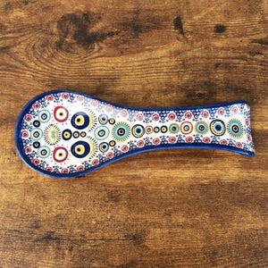 May include: A ceramic spoon rest with a blue rim and a colorful pattern of circles and floral designs. The spoon rest is white with red, yellow, blue, and black accents. The spoon rest is on a wooden surface.