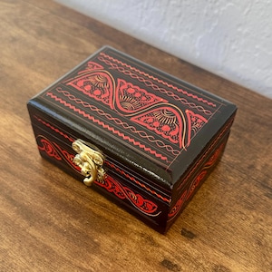 May include: A small, rectangular wooden box with a black finish and intricate red designs. The box features a gold-colored clasp and decorative patterns on the top and sides. The box is sitting on a wooden surface.