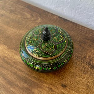 May include: A round, lidded decorative box with a black finial. The box is painted with a green and gold floral design on a black background. The box sits on a wooden surface.