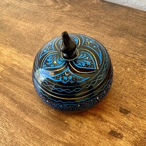 May include: A round, black decorative box with a lid. The box is adorned with blue and gold floral patterns. The lid has a small, black knob. The box is sitting on a wooden surface.