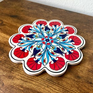 May include: A white, scalloped-edge ceramic coaster with a vibrant floral design. The coaster features red, blue, and navy floral patterns on a white background. The coaster is approximately 6 inches in diameter.